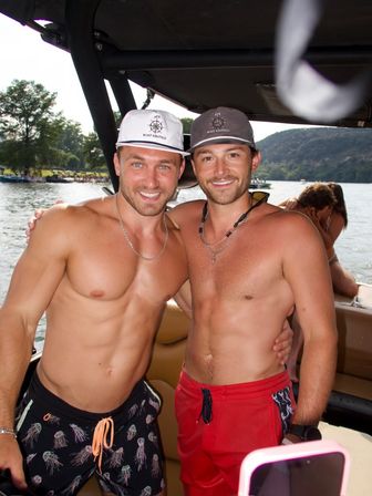 Two shirtless men smiling together on a motorboat during a sunny lake day, wearing caps and swim trunks with wooded shoreline and hills in the background.