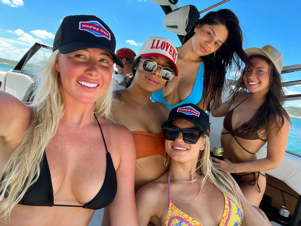 Five friends in bikinis and caps smiling on a motorboat near turquoise coastal waters on a sunny summer day