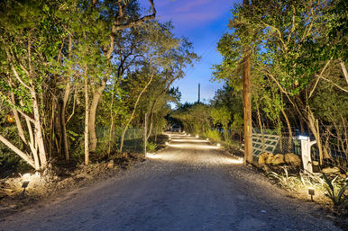 Tree-lined rural gravel driveway at dusk, warmly lit by low landscape lights along a fence and mailbox, leading toward a distant home.