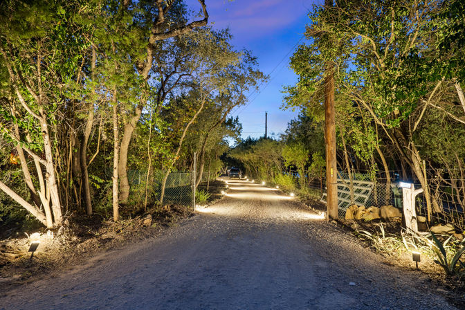 Tree-lined rural gravel driveway at dusk, warmly lit by low landscape lights along a fence and mailbox, leading toward a distant home.