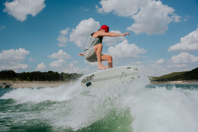 Wakesurfer in a red cap and gray life vest launching off a white board over a green lake wake, splashing water under a bright blue sky with puffy clouds and a tree-lined shoreline.