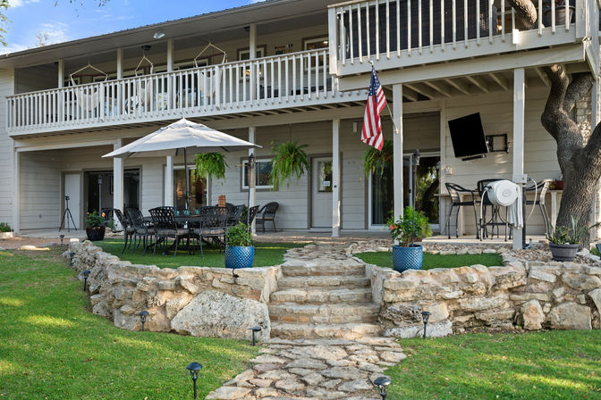Two-story backyard patio with stone steps and rock retaining wall leading to an outdoor dining area under a white umbrella, black metal table and chairs, hanging ferns, blue potted plants, American flag on the porch, upper balcony with white railing, wall-mounted TV, bar stools, and green lawn with pathway lights.