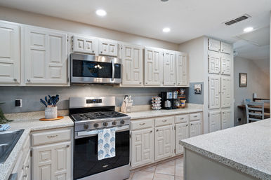 Bright residential kitchen with white paneled cabinets, stainless steel microwave and gas range, speckled quartz countertops, built-in coffee station, recessed lighting and a small breakfast nook visible in the background.