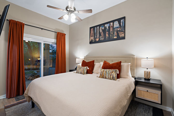 Cozy Austin-themed bedroom with king bed and tufted headboard, rust-colored curtains framing a sliding glass door, patterned throw pillows, ceiling fan, and bedside lamps on black nightstands.