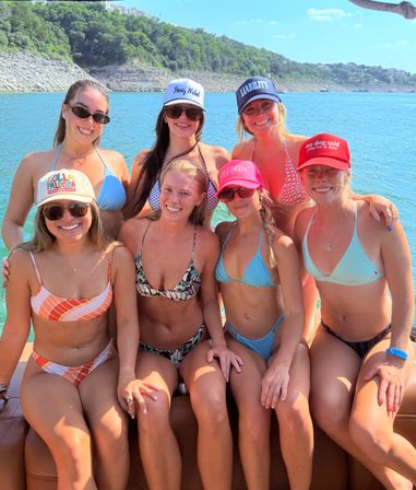 Seven friends in colorful bikinis and caps smiling and posing on a boat over a sunny turquoise lake with a tree-covered rocky shoreline