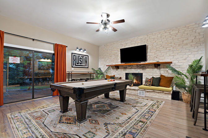 Inviting game room with dark wood pool table on a patterned rug, stone fireplace with TV above, built-in bench with colorful pillows, potted plants, sliding glass door to backyard, warm lighting and orange curtains.