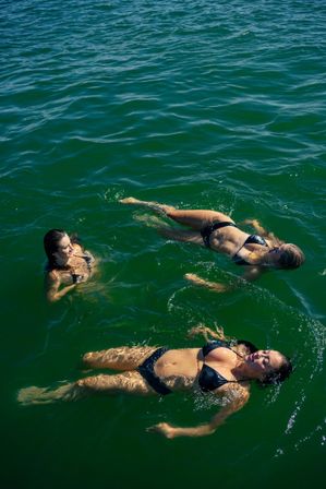 Three women in black bikinis floating and relaxing in sunlit emerald-green lake water—casual summer swim scene