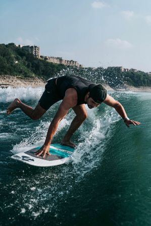 Surfer crouches on a turquoise surfboard carving a green wave near a rocky coastal shoreline under a clear blue sky.