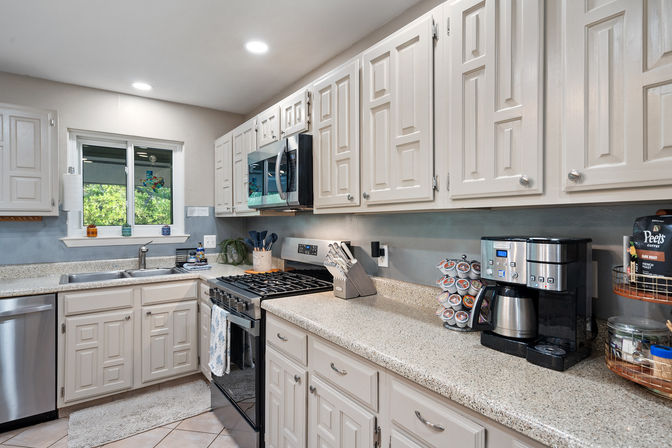 Bright, cozy residential kitchen with white paneled cabinets, speckled beige countertops, stainless-steel gas range and microwave, sink under a window with a green yard view, and a countertop coffee station.
