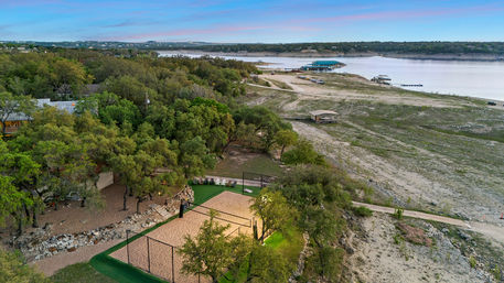 Aerial view of a sandy lakeside volleyball court tucked among oak trees, with rocky shoreline, docks and a calm blue-pink sky over the lake.