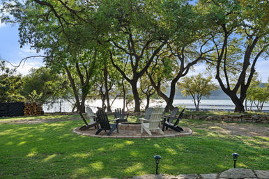 Inviting lakefront backyard with a circular stone fire pit surrounded by Adirondack chairs under shady trees, stacked firewood and a calm lake with a dock in the background.