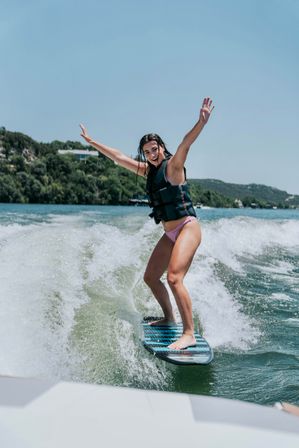 Smiling woman wakesurfing on a sunny lake, balancing on a blue-striped board with arms raised, wearing a life vest and pink bikini bottoms as the boat wake and tree-lined shoreline roll by.