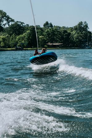 Two riders on a blue towable tube catch air over a boat wake on a sunny lake with a tree-lined shoreline