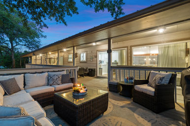 Cozy covered backyard deck at dusk with wicker sectional and armchair, cushioned pillows, glass-top coffee table with candles, hammock, wooden railing and sliding glass doors opening to a warmly lit home.