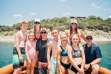 Smiling multigenerational family and kids in swimsuits posing on a boat at a sunny lake with a tree-lined shoreline and a house on the hill in the background.