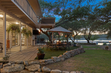 Cozy lakeside backyard patio at dusk with umbrella-shaded dining table and wrought-iron chairs, hanging ferns, stone border and second-story balcony overlooking a calm lake