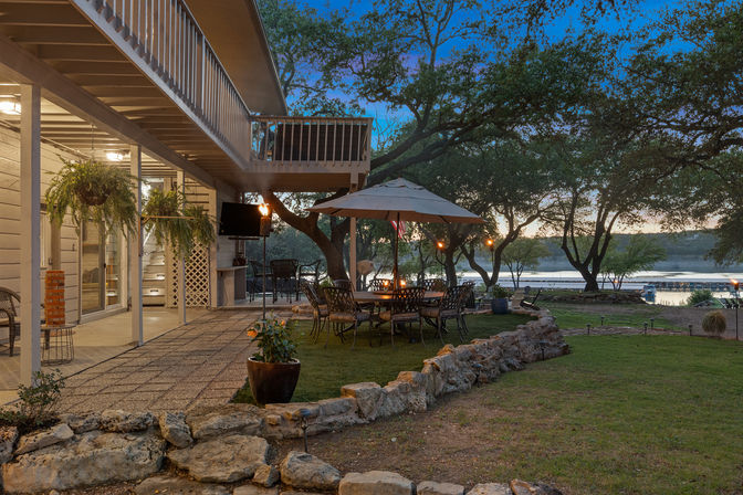 Cozy lakeside backyard patio at dusk with umbrella-shaded dining table and wrought-iron chairs, hanging ferns, stone border and second-story balcony overlooking a calm lake