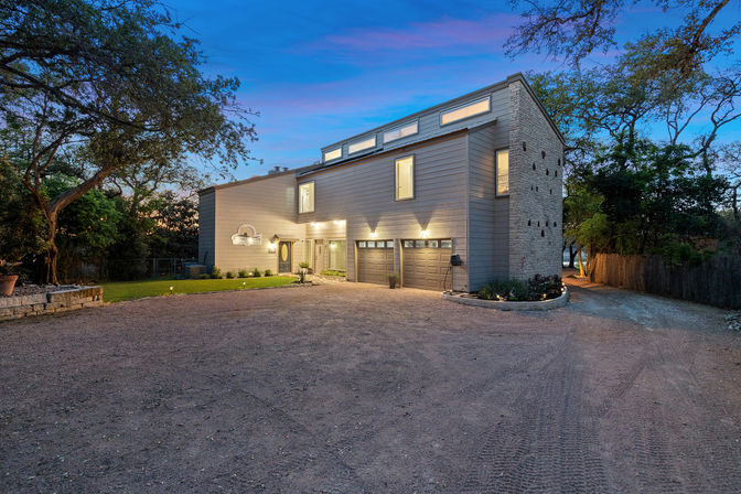 Contemporary two-story home with three-car garage, warm exterior lighting at twilight, gravel driveway, mature oak trees and a glimpse of water in the background.