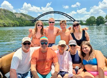 Smiling group of friends and family on a boat in swimsuits, posing on a sunny summer day with the arched Pennybacker (360) Bridge over Lady Bird Lake in Austin, Texas in the background.