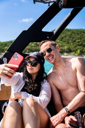 Smiling shirtless man and woman in sunglasses taking a red-phone selfie on a sunny summer boat ride over turquoise water with a green shoreline, both holding canned drinks