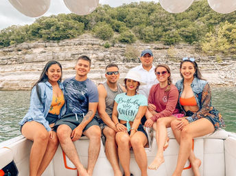 Seven friends on a boat enjoying a summer lake day with rocky cliffs and wooded shoreline in the background, casual swimwear, sun hat and balloons overhead.