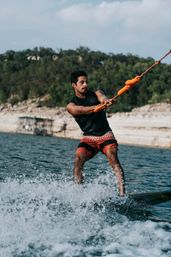 Wakeboarder on a lake gripping an orange tow rope as spray kicks up; wearing a black life vest and red board shorts with rocky shoreline and tree-covered hills in the background.
