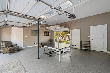 Spacious residential garage converted into a game room with a ping pong table at center, dartboard on the wall, leather armchair, open garage doors showing a suburban backyard with trees, and a clean epoxy-coated floor — ready for a match.