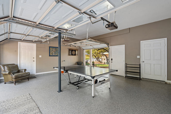 Spacious residential garage converted into a game room with a ping pong table at center, dartboard on the wall, leather armchair, open garage doors showing a suburban backyard with trees, and a clean epoxy-coated floor — ready for a match.