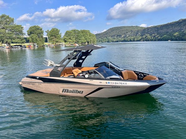Sleek tan-and-black wakeboard boat with a wake tower floating on a calm lake on a sunny day, with tree-lined shores and green hills in the background.
