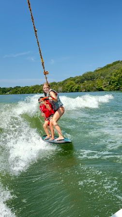 Two people tandem wakeboarding on a sunny lake — a smiling young woman and child in life jackets hold the tow rope on a blue board, riding the green wake with a tree-lined shoreline.