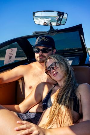 Smiling couple in sunglasses and swimwear relaxing in a speedboat on a sunny lake, rearview mirror reflecting the boat’s wake and tow ropes under a clear blue sky