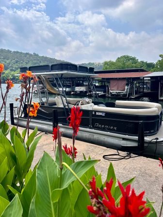 Pontoon boat moored at a lakeside dock and marina with bright red tropical flowers in the foreground and green hills under a partly cloudy sky.