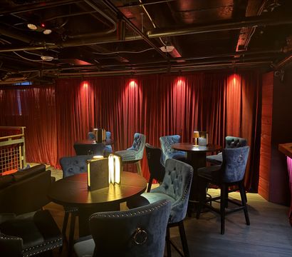 Cozy velvet-draped cocktail lounge interior with red curtains, tufted blue velvet chairs, round dark-wood tables, small table lamps and warm mood lighting under exposed black ceiling pipes.