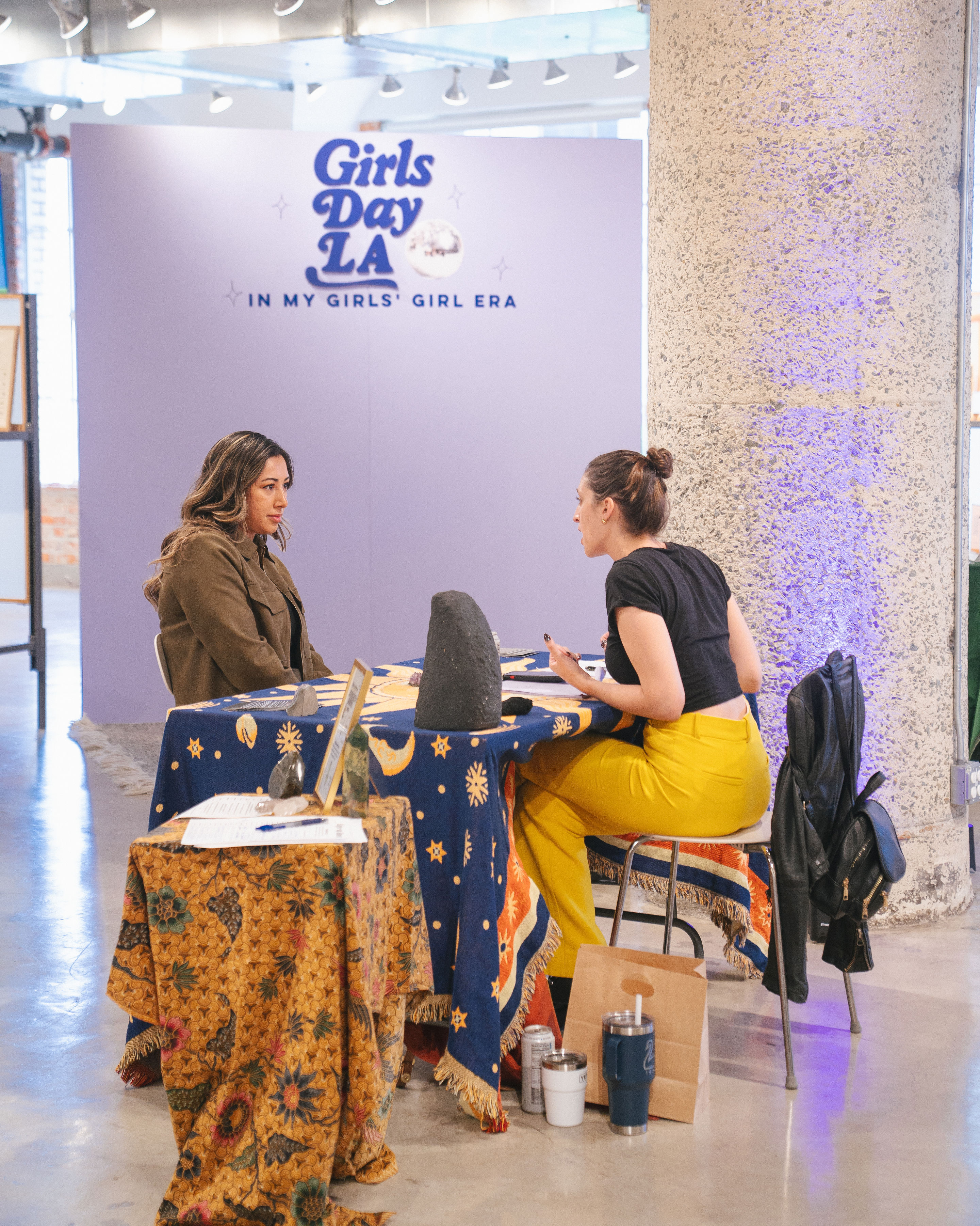 Two women seated across a star-and-moon covered table of crystals and stones, chatting at an indoor Girls Day LA pop-up with a lavender backdrop and textured concrete column.