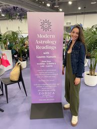 Smiling woman poses beside a tall purple banner for Modern Astrology Readings at a lively indoor expo booth, surrounded by potted palms and hanging lavender florals.