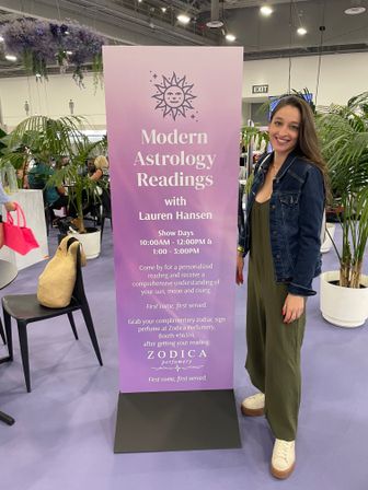 Smiling woman poses beside a tall purple banner for Modern Astrology Readings at a lively indoor expo booth, surrounded by potted palms and hanging lavender florals.
