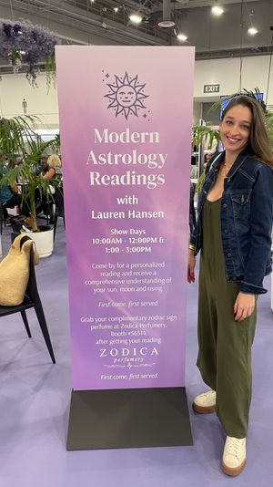 Smiling woman poses beside a tall purple banner for Modern Astrology Readings at a lively indoor expo booth, surrounded by potted palms and hanging lavender florals.
