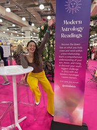 Smiling woman in yellow pants raises her arm at an indoor expo astrology booth next to a tall purple Modern Astrology Readings banner on a bright pink trade-show floor with floral decor and clear bar stools.