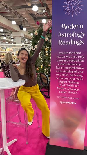 Smiling woman in yellow pants raises her arm at an indoor expo astrology booth next to a tall purple Modern Astrology Readings banner on a bright pink trade-show floor with floral decor and clear bar stools.