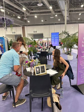 Three people chatting at a round table inside a lively convention center lounge with potted palms, a floral centerpiece, booths and an events schedule screen in the background.