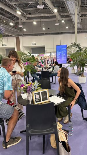 Three people chatting at a round table inside a lively convention center lounge with potted palms, a floral centerpiece, booths and an events schedule screen in the background.