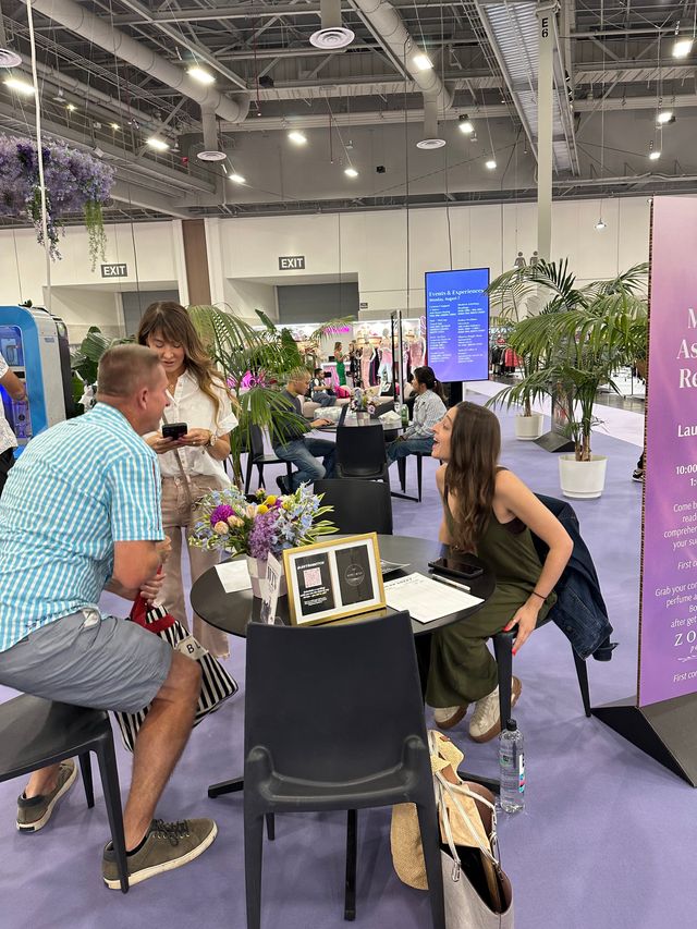 Three people chatting at a round table inside a lively convention center lounge with potted palms, a floral centerpiece, booths and an events schedule screen in the background.