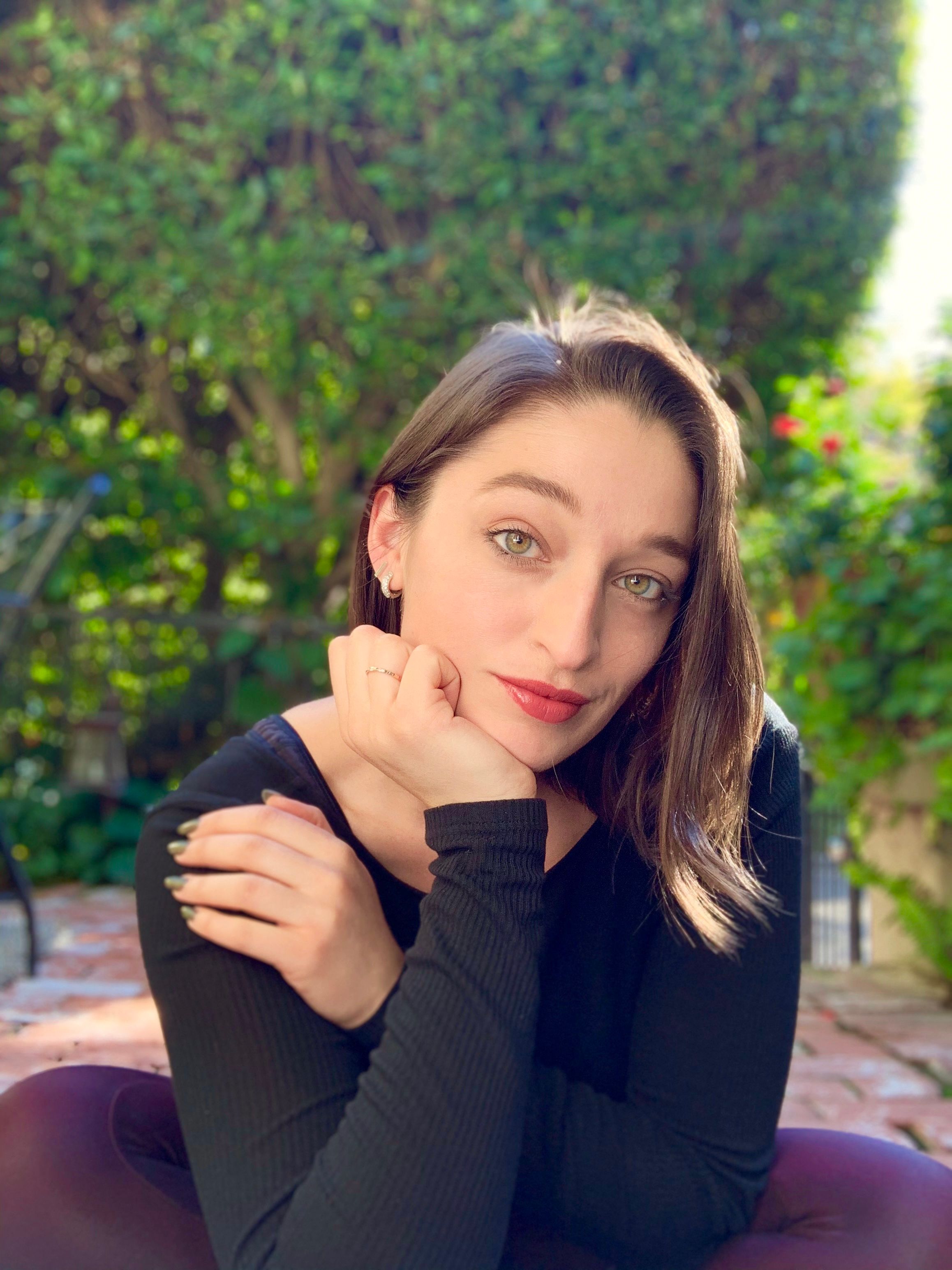 Sunlit close-up portrait of a woman with green eyes and red lipstick resting her chin on her hand in a leafy backyard garden with a blurred hedge and colorful flowers