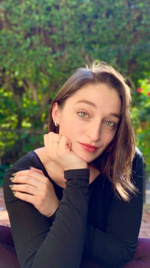 Sunlit close-up portrait of a woman with green eyes and red lipstick resting her chin on her hand in a leafy backyard garden with a blurred hedge and colorful flowers