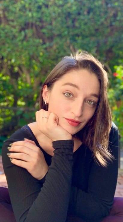 Sunlit close-up portrait of a woman with green eyes and red lipstick resting her chin on her hand in a leafy backyard garden with a blurred hedge and colorful flowers