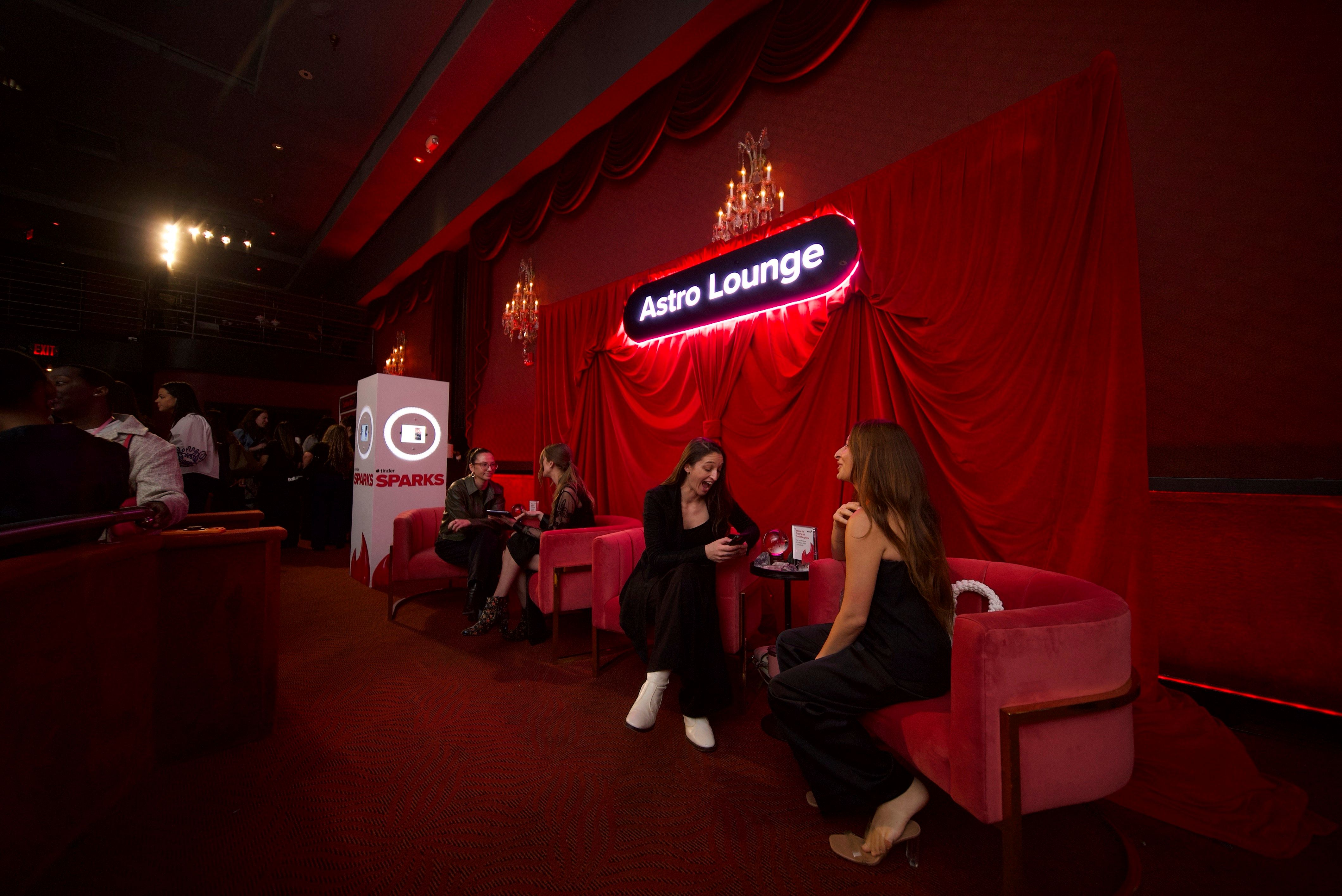 Red velvet lounge interior with draped curtains, chandeliers and a glowing neon sign above pink velvet chairs where groups chat and relax at a lively cocktail event
