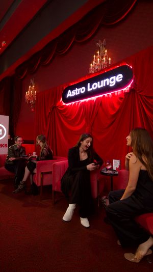 Red velvet lounge interior with draped curtains, chandeliers and a glowing neon sign above pink velvet chairs where groups chat and relax at a lively cocktail event