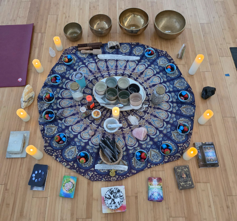 Mandala cloth altar on a wooden studio floor with singing bowls, LED candles, crystals, small cups and colorful stones, tarot/oracle decks and ritual tools arranged for a meditation or healing circle in a yoga/wellness space.