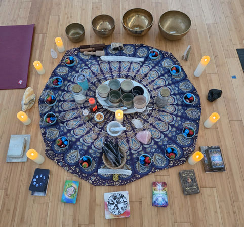 Mandala cloth altar on a wooden studio floor with singing bowls, LED candles, crystals, small cups and colorful stones, tarot/oracle decks and ritual tools arranged for a meditation or healing circle in a yoga/wellness space.