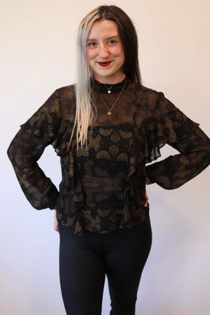 Studio portrait of a person with half-black, half-platinum hair wearing a sheer black blouse with gold paisley print and black leggings, hands on hips and smiling against a white background.